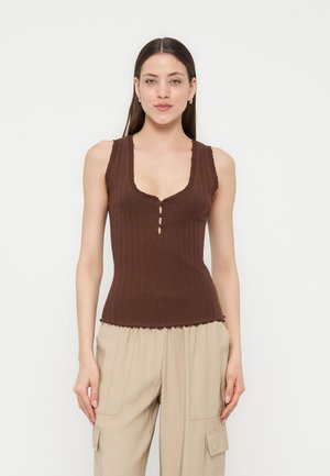 Γυναίκα με μακριά σκούρα μαλλιά, που φοράει αμάνικο καφέ ριμπ τοπ με τρία μικρά κουμπιά μπροστά και μπεζ cargo παντελόνια, στέκεται μπροστά σε λευκό φόντο.