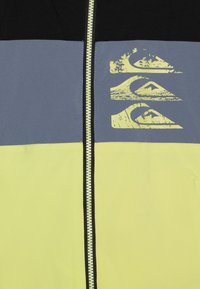 Jopica z zadrgo v barvah blokov, ki vsebuje črne, sive in neonsko rumene panele ter teksturirano logotipno zasnovo na prsih. Gladka tekstura materiala.