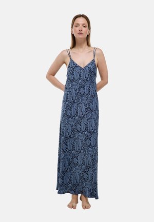 Femme debout pieds nus vêtue d'une robe longue sans manches bleue à motif floral avec fines bretelles doubles sur un fond uni.