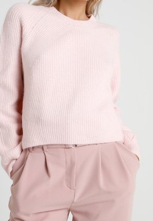 Woman wearing a light pink ribbed knit sweater with long sleeves tucked into high-waisted, pleated blush pink trousers with hands in pockets.