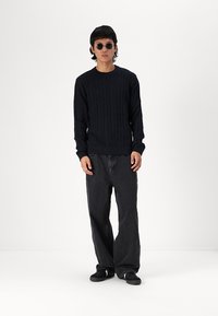 Marine kabelstrikket genser, avslappet passform, rund hals. Kombinert med svarte vidbenede jeans og svarte sko. Modell med runde solbriller.