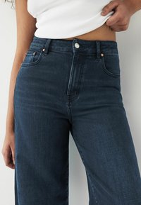 Mørke denim bukser med vid ben og høj talje, der har en frontknaplukning, to sidelommer og en glat overflade. Fremhævede sømme.