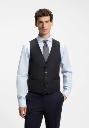 Mann mit lockigem Haar, der ein hellblaues Hemd, gemusterte Krawatte und eine schwarze, zugeknöpfte Weste trägt, steht mit einer Hand in der Tasche.