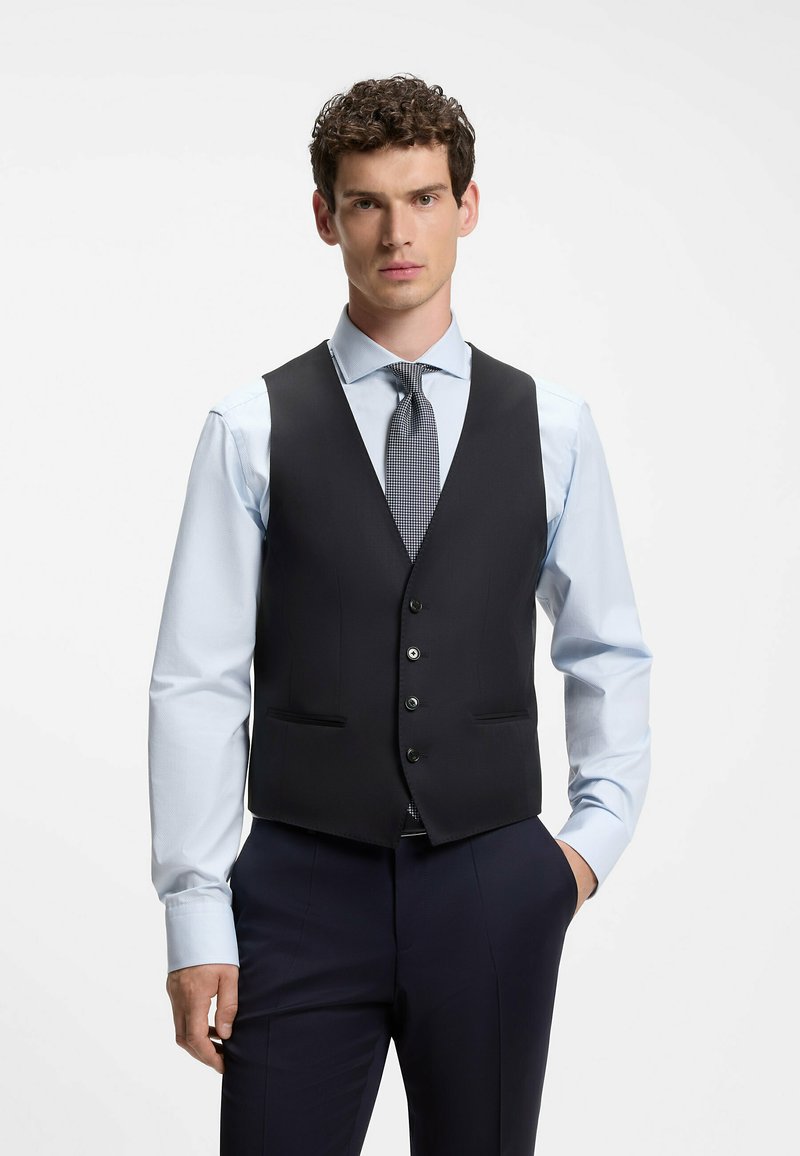 Homme aux cheveux bouclés portant une chemise bleu clair, une cravate à motifs et un gilet noir boutonné, debout avec une main dans la poche.