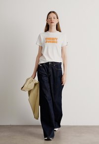 Biała koszulka z pomarańczowym napisem "Tommy Jeans", w połączeniu z ciemnoniebieskimi szerokimi dżinsami. Model trzyma beżową kurtkę na jednym ramieniu.