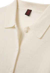 Polo shirt lavorato a maglia color crema con colletto, tre bottoni bianchi e etichetta nera con testo rosso "STEFANEL."