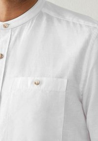 Bílá lněná košile s mandarin límečkem, zapínáním na knoflíky v přední části s béžovými knoflíky a jednou náprsní kapsou. Hladká textura a minimalistický design.