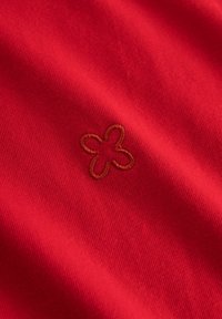 Roter Baumwollstoff mit glatter Textur, der in der Mitte ein braunes, gesticktes Blumendesign aufweist. Schlichte und minimalistische Ästhetik.