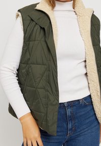 Grüne gesteppte Weste mit einem beigen Sherpa-Innenfutter, ausgestattet mit einem Reißverschluss auf der Vorderseite und seitlichen Taschen, kombiniert mit einem taillierten weißen Rippenrollkragen.