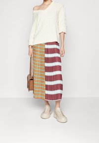 Sweater crème texturé à épaules dénudées associé à une jupe midi plissée bicolore avec des rayures marron, orange et bleue ; baskets beiges.