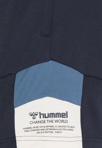 Hummel-logo ja slogan "Muuta maailmaa" tummansinisen, sinisen ja valkoisen kankaan päällä, jossa on tekstiä mukavuudesta ja optimismista aktiivisessa elämäntavassa.