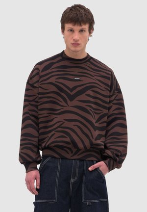 Junger Mann trägt ein braun-schwarz gestreiftes Sweatshirt mit Zebramuster und kleinem Brustlogo, kombiniert mit dunkelblauen Jeans mit weißen Nähten.