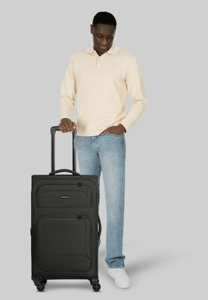 Mann in beigem Langarmhemd und Jeans steht neben einem großen schwarzen Rollkoffer mit ausgezogenem Griff.