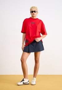 Raudona per didelė T-marškiniai su juodu grafiniu spaudiniu, derinama su tamsiomis džinsinėmis sijonais. Modelis dėvi juodus akinius nuo saulės ir baltus „Nike“ sportbačius.