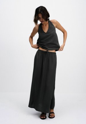 Frau justiert dunkles ärmelloses Neckholder-Top und trägt passenden langen Rock sowie schwarze Sandalen vor schlichtem hellem Hintergrund.