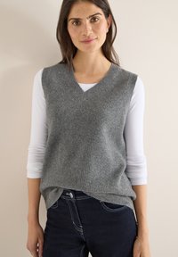 Maglione smanicato grigio a maglia con scollo a V, indossato sopra una camicia bianca a maniche lunghe, abbinato a jeans scuri con cuciture a contrasto.