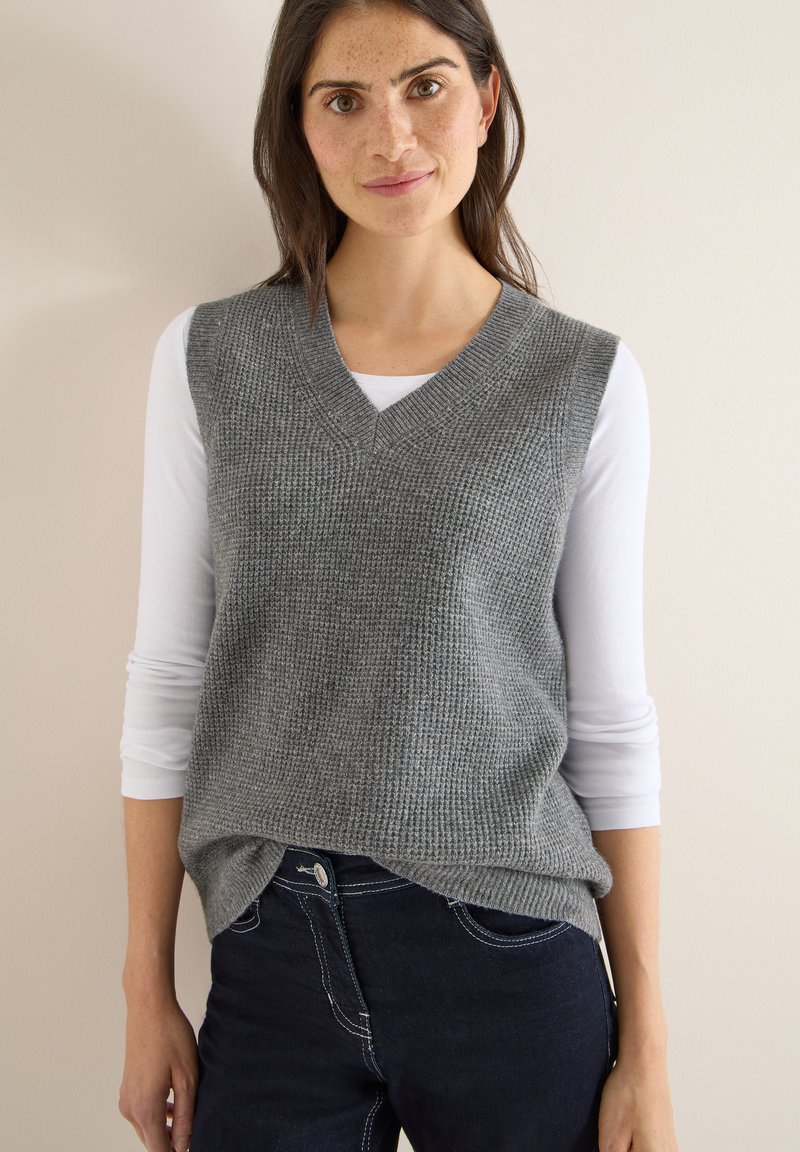 Maglione smanicato grigio a maglia con scollo a V, indossato sopra una camicia bianca a maniche lunghe, abbinato a jeans scuri con cuciture a contrasto.