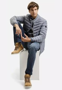 Szara kurtka puchowa z kołnierzem stójkowym, dżinsy i brązowe buty z białą podeszwą, siedząca na białym sześcianie na jasnym tle.