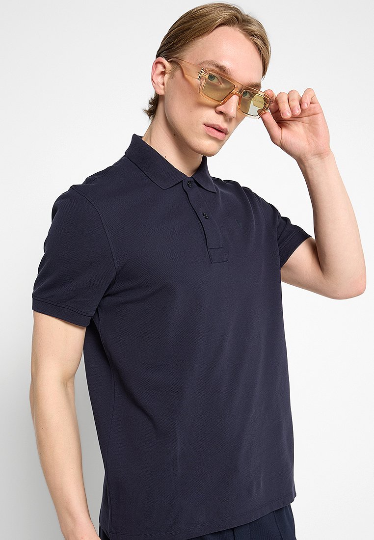 Marineblaues Poloshirt, kurze Ärmel, strukturierten Stoff, mit drei Knöpfen, getragen mit klaren Sonnenbrillen, Modell platziert die Hand auf der Brille.