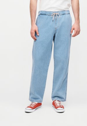 Mann trägt hellblaue Jeans mit elastischem Bund und Kordelzug sowie rote Canvas-Sneakers, steht vor weißem Hintergrund.