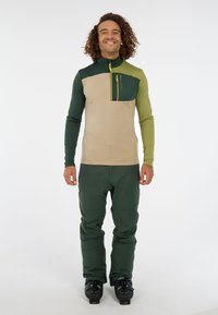 Heren buitenjack in beige en groen met een halvemaanrits, lange mouwen en een borstzak; gecombineerd met groene skibroek en zwarte laarzen.