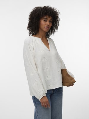 Femme aux cheveux bouclés porte un chemisier blanc à manches longues avec des détails en broderie anglaise et un jean bleu, tenant une pochette en daim marron, sur fond blanc uni.