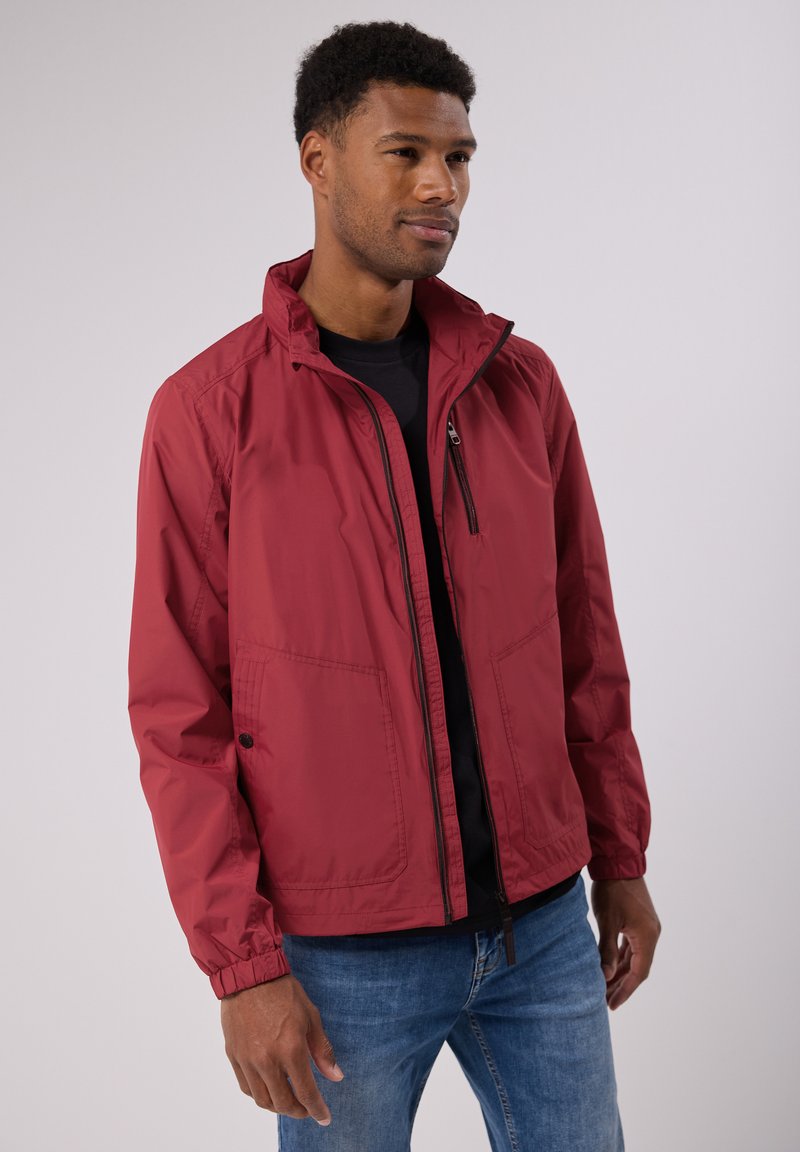 Veste légère rouge avec un col haut, fermeture éclair et poignets élastiques. Comprend deux poches avant et une texture lisse, résistante à l'eau.