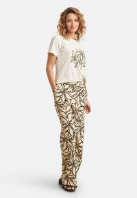 Witte t-shirt met zwarte bloemenprint, gecombineerd met beige broek met een gedurfde zwarte bloemenpatroon. Het model staat in een ontspannen houding.