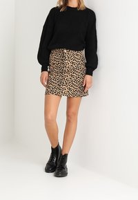 Schwarzer Wollpullover mit überschnittenen Schultern, kombiniert mit einem beigen Minirock im Leopardenmuster mit vorderen Knöpfen. Schwarze Schnürstiefel bis zum Knöchel.