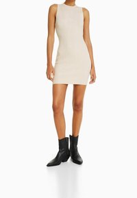 Robe en maille côtelée beige avec une silhouette ajustée et sans manches, associée à des bottes chelsea en cuir noir avec un bout pointu.