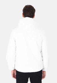 Uomo con capelli castani corti, che indossa una giacca bianca con cappuccio e pantaloni neri, in piedi con la schiena rivolta verso uno sfondo neutro.