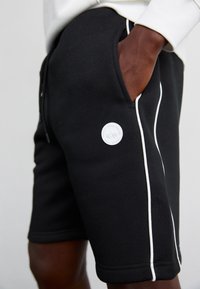 Short noir avec passepoil blanc et petit logo rond blanc sur la cuisse, porté avec un haut à manches longues de couleur claire, main dans la poche.