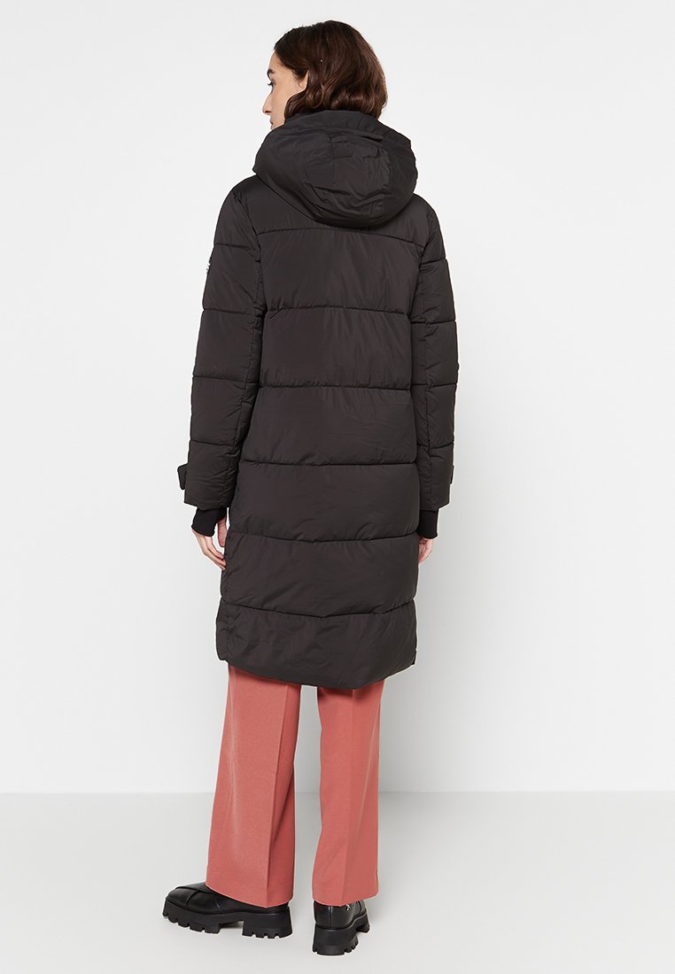 Schwarze Steppjacke mit horizontalen Gesteppten Abschnitten, Kapuze und langen Ärmeln, kombiniert mit hellrosanen Hosen und klobigen schwarzen Stiefeln.