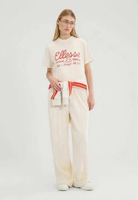Crèmekleurig T-shirt met rood "Ellesse" logo, gecombineerd met losse, crèmekleurige broek met zijnaden en geaccentueerd met een riem met rode strepen.