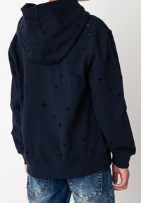 Sudadera azul marina con acabado texturizado, que presenta múltiples agujeros desgastados. El diseño incluye un bolsillo frontal tipo pouch y una capucha con cordón.