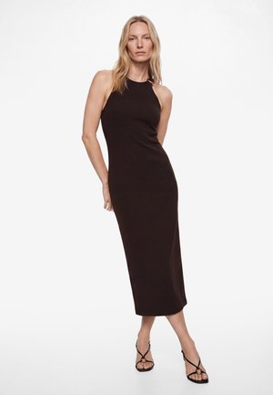 Femme blonde portant une robe midi noire cintrée sans manches et des sandales à talons hauts noires à brides, debout sur fond blanc.