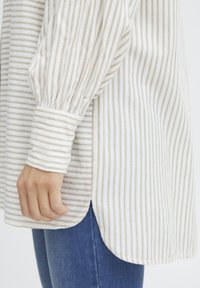 Striped long-sleeve shirt in white and light beige, featuring a rounded hem and gathered cuffs. Soft fabric with a relaxed fit.