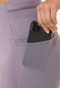 Lila Leggings mit einer seitlichen Tasche für ein Smartphone, gefertigt aus dehnbarem Stoff mit einer glatten Textur und genähten Akzenten.