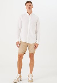 Weiße Leinenbluse mit Knopfverschluss, beiger, umgeschlagener Shorts und beigen Sneakers mit weißen Akzenten, die vor einem schlichten Hintergrund stehen.