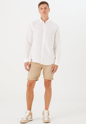 Camisa blanca de lino con cierre de botones, pantalones cortos beige enrollados y zapatillas beige con acentos blancos, de pie contra un fondo simple.