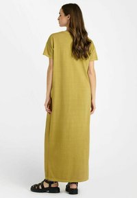 Maxi-jurk in olijfgroen, gemaakt van lichtgewicht stof, met korte mouwen en zijsplitten voor bewegingsgemak. Zwarte sandaaltjes met bandjes maken de look compleet.