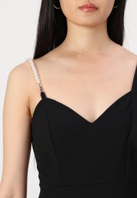 Sort kjole med en struktureret v-halsudskæring, prydet med perle detaljer på stropperne, glat stof og en tætsiddende silhuet.