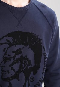 Felpa blu navy con scollo rotondo, caratterizzata da una grande grafica nera di un profilo laterale con mohawk e la scritta "DIESEL". Tessuto testurizzato.