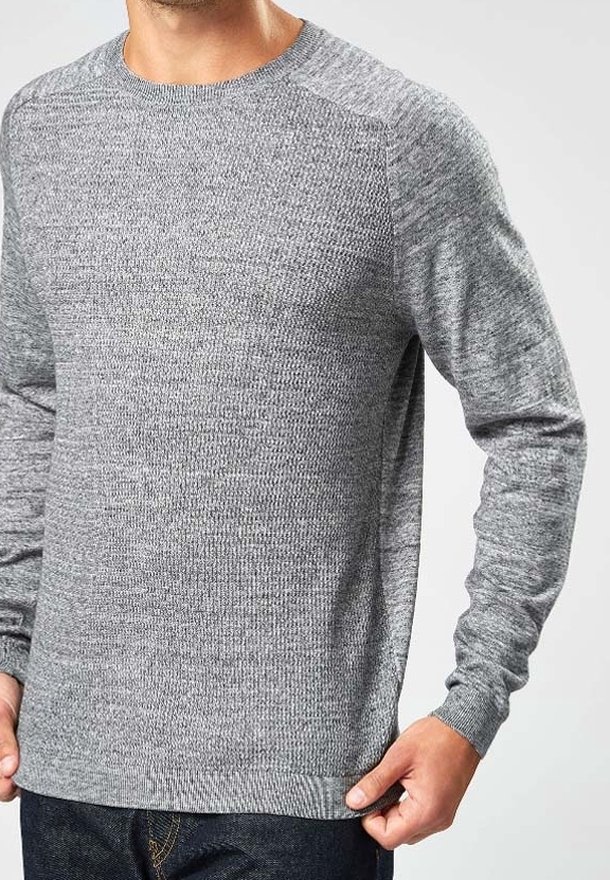 Homme portant un pull tricoté gris clair à manches longues avec un col rond, tenant l'ourlet d'une main.