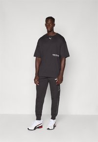 Juodas medvilninis oversized t-shirt su baltu Puma logotipu; juodos sportinės kelnės su baltu apvadu ir Puma ženklinimu; baltos ir raudonos sportinės bateliai.