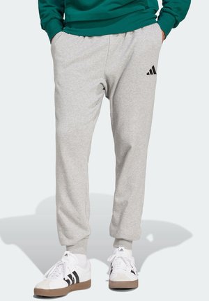 Osoba nosząca jasnoszare spodnie dresowe Adidas z rękami w kieszeniach oraz białe sneakersy Adidas z czarnymi paskami.