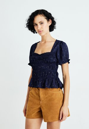 Marineblauwe blouse met smockdetail, vierkante halslijn, korte pofmouwen, gouden stippenpatronen en een peplumrand, gecombineerd met tan suede shorts.