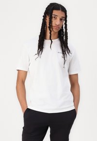 Camiseta blanca de manga corta de algodón con un logo en el lado izquierdo del pecho. Textura suave, corte estándar, combinada con pantalones negros.