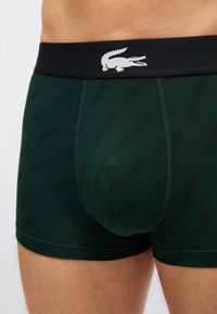 Σκούρα πράσινα μπόξερ με επαναλαμβανόμενο μοτίβο κειμένου "Lacoste" και μαύρη ζώνη μέσης με λευκό λογότυπο κροκοδείλου, φορεμένα από ένα άτομο.