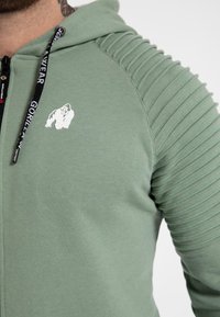 Sudadera con cremallera verde claro, con detalles acanalados en los hombros, logo de gorila blanco y cordones negros que presentan el texto de la marca. Textura de algodón suave.